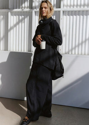 Person wearing a black outfit holding a white cup against a white wall with shadows.