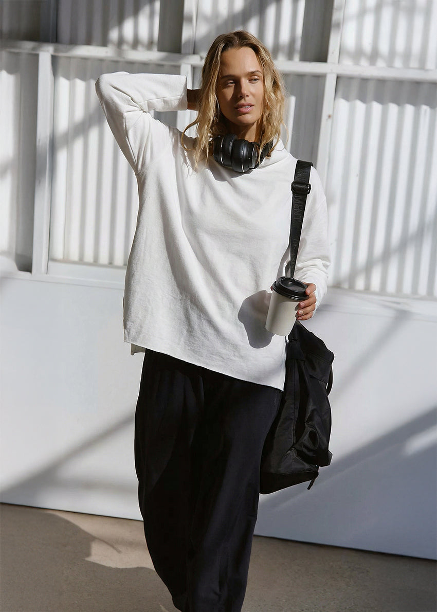 Person wearing a white shirt and black pants, holding a coffee cup and umbrella, standing against a white wall.