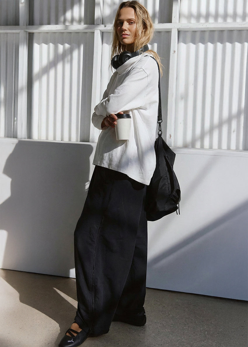 Person wearing a white shirt and black pants with a black bag against a white wall.