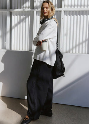 Person wearing a white shirt and black pants with a black bag against a white wall.