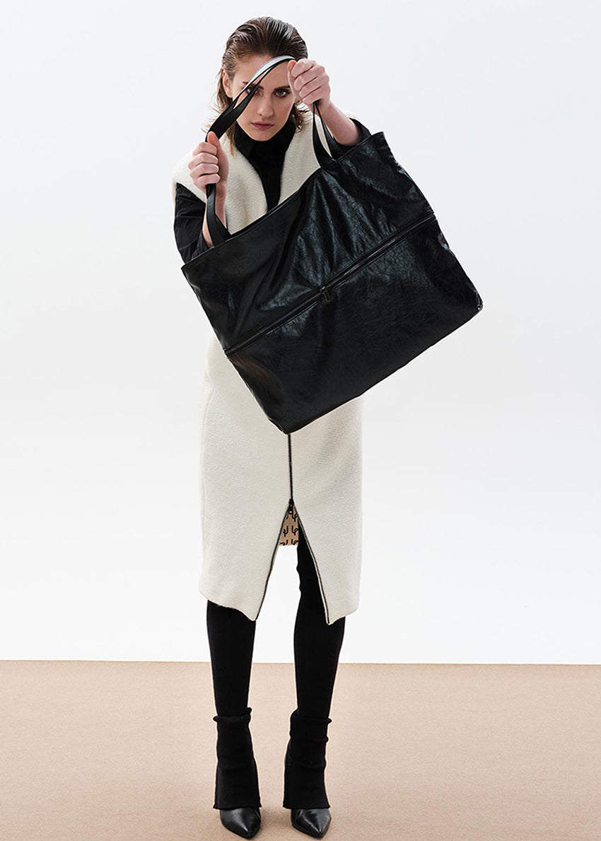 A person holding an oversized black tote bag with a zippered top and shoulder straps.