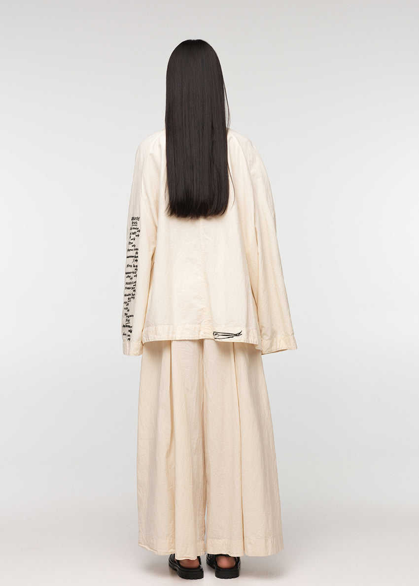 A model wearing a beige oversized jacket with an open collar, featuring four front pockets and closures with buttons. The jacket has a relaxed fit and the model is also wearing light-colored pants.