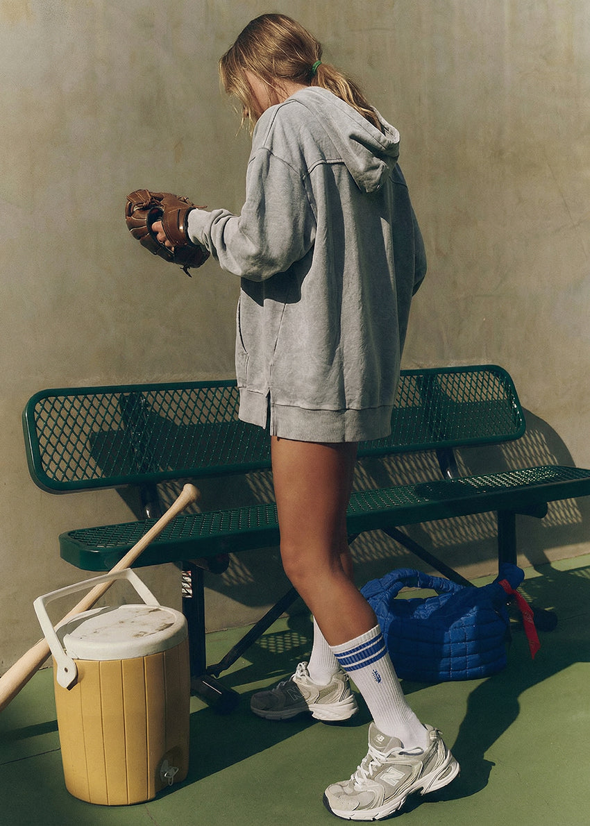 Woman sitting on a green bench holding a baseball bat, wearing a gray hoodie and yellow shorts.