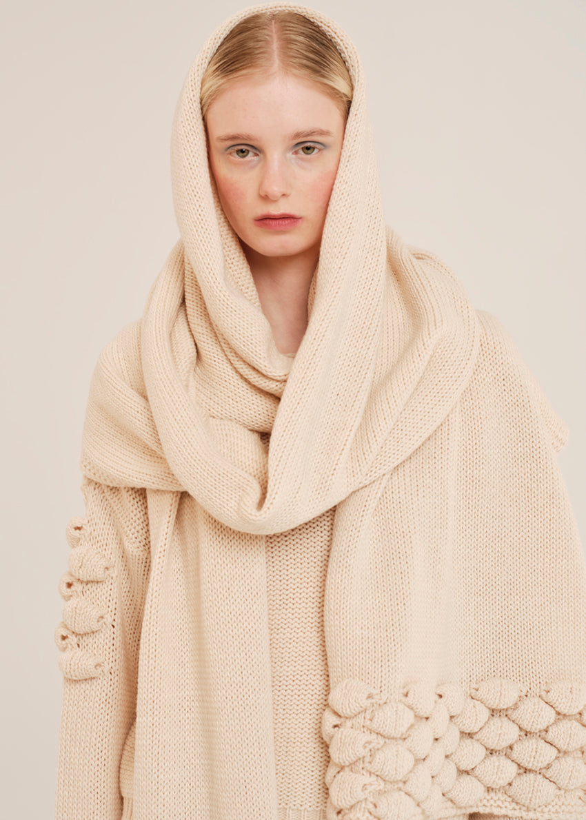 Woman wearing a beige knitted sweater and scarf against a plain background