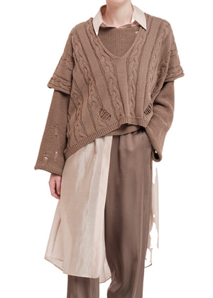 Person wearing a brown cable knit vest over a white shirt with beige pants on a white background