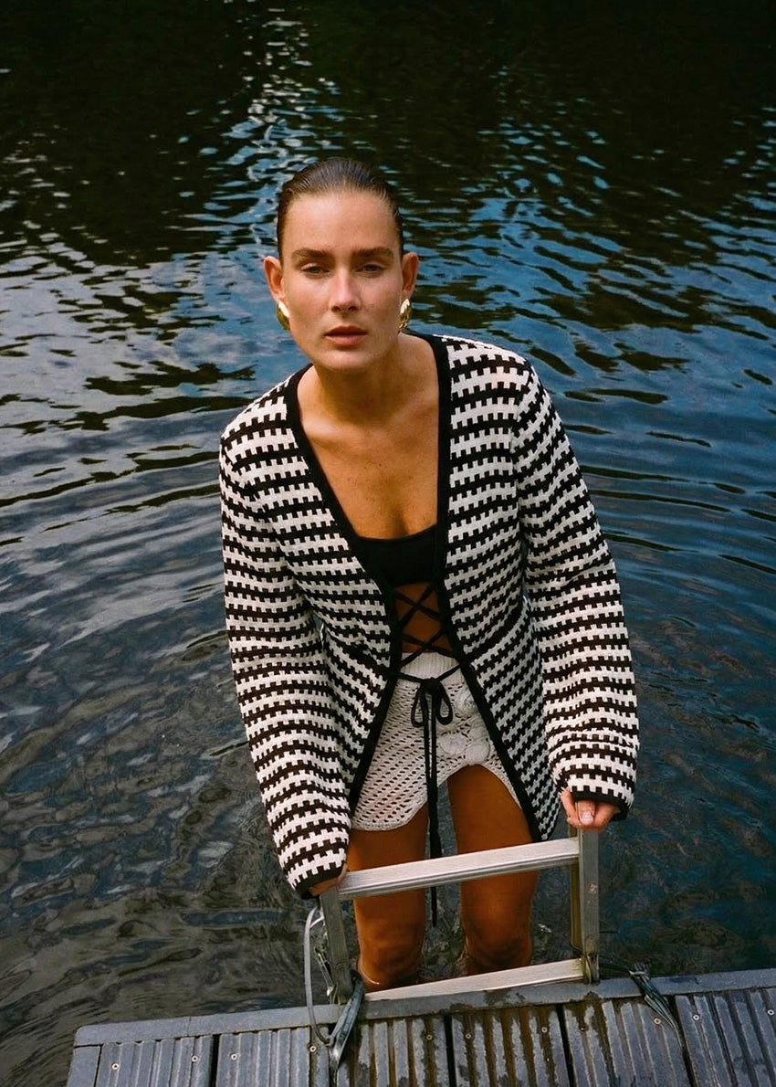 Woman standing on a dock by water wearing a black and white patterned cardigan.