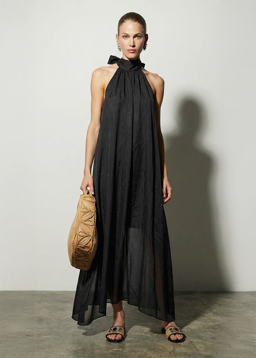 Woman wearing a black halter neck dress holding a beige bag against a plain background