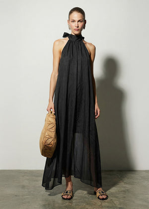 Woman wearing a black halter neck dress holding a beige bag against a plain background