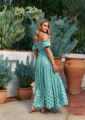 A woman standing in a garden wearing a green off-shoulder maxi dress with a geometric pattern.
