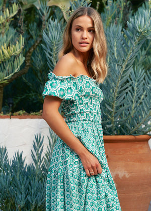 A woman standing in a garden wearing a green off-shoulder maxi dress with a geometric pattern.