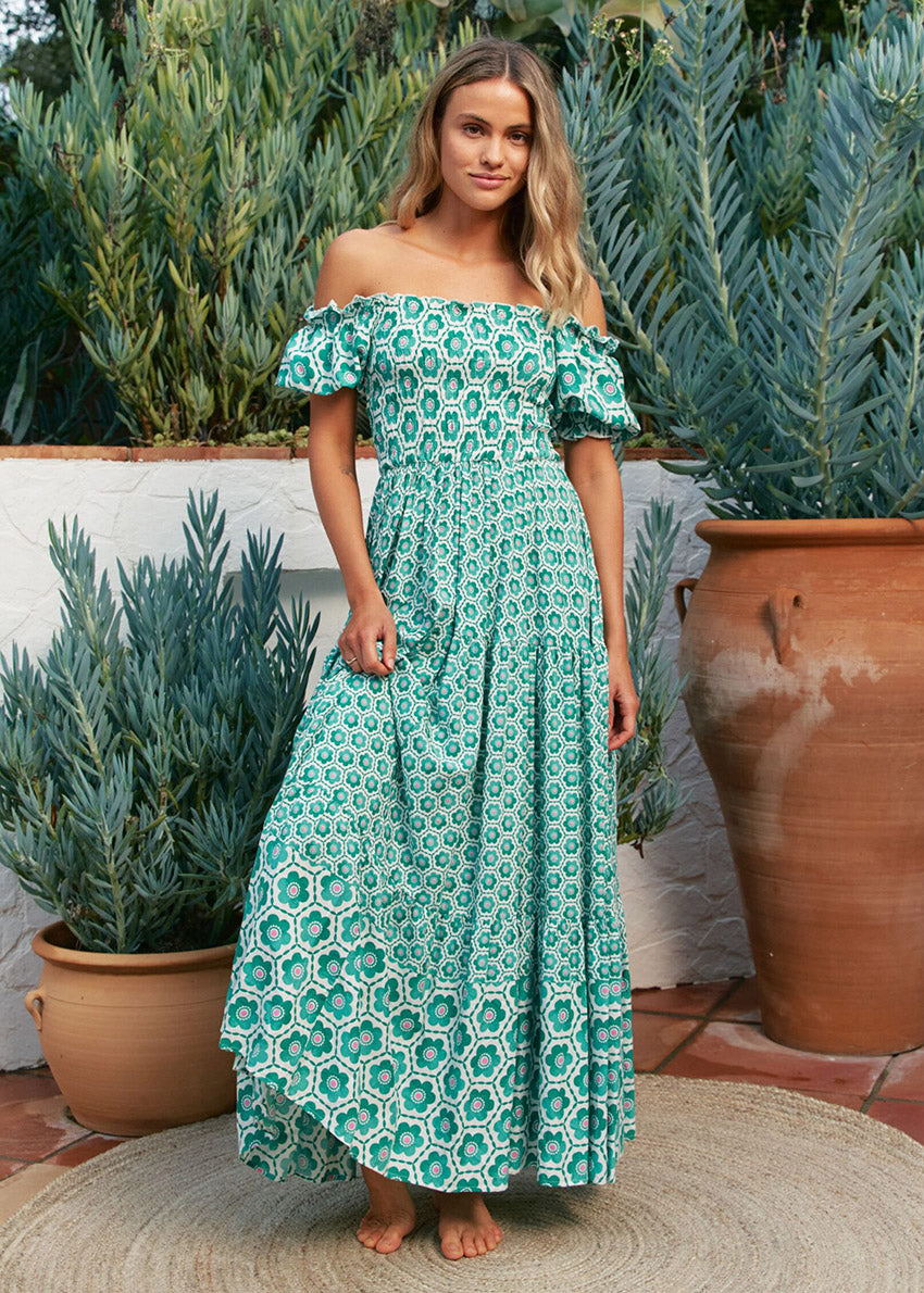 A woman standing in a garden wearing a green off-shoulder maxi dress with a geometric pattern.