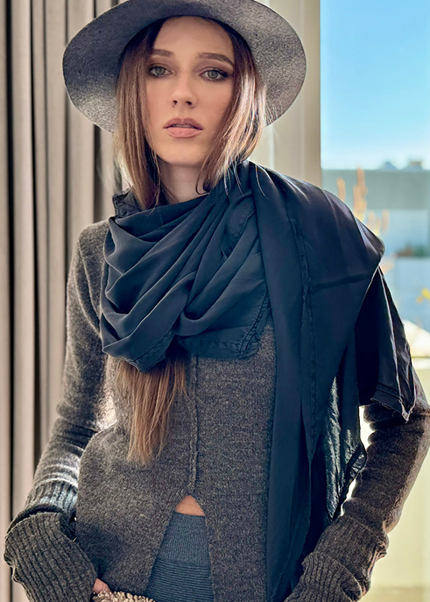 A woman wearing a gray sweater and a navy blue scarf with a wide slit, paired with a gray hat, posing indoors.