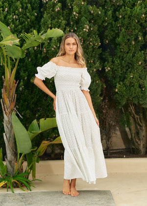 A woman wearing an off-the-shoulder white maxi dress with short puff sleeves and a tiered design, standing near greenery.
