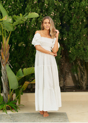 A woman wearing an off-the-shoulder white maxi dress with short puff sleeves and a tiered design, standing near greenery.