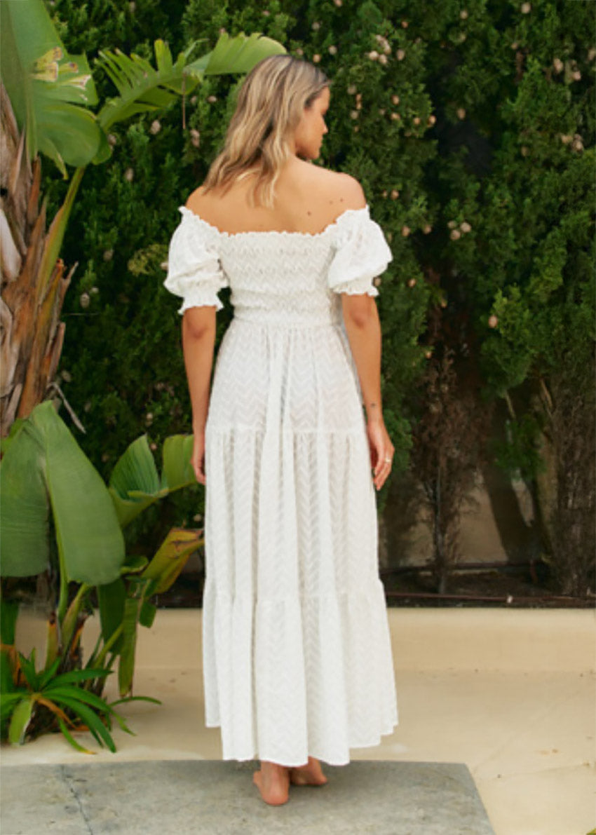 A woman wearing an off-the-shoulder white maxi dress with short puff sleeves and a tiered design, standing near greenery.