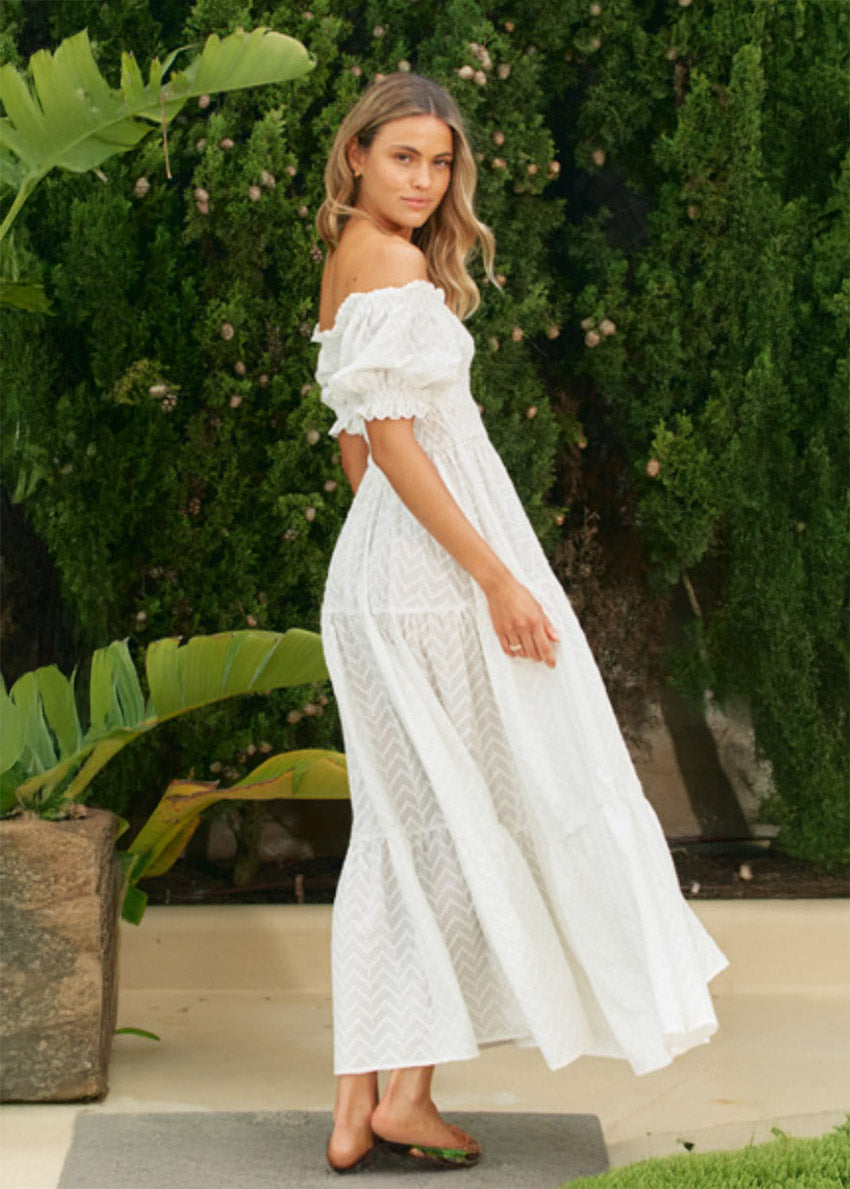 A woman wearing an off-the-shoulder white maxi dress with short puff sleeves and a tiered design, standing near greenery.