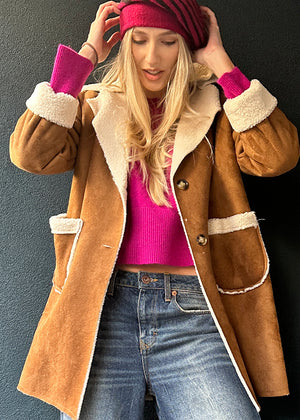 Person wearing a brown coat with white fur lining, pink sweater, and blue jeans against a gray background.