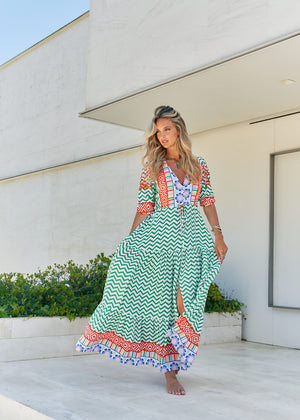 A woman standing in front of a building entrance wearing a floral and geometric patterned maxi dress with a V-neckline and billowy sleeves.