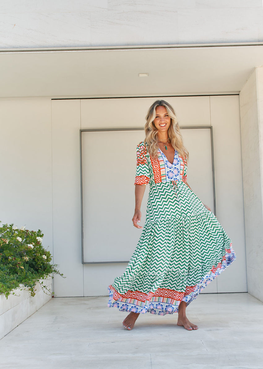 A woman standing in front of a building entrance wearing a floral and geometric patterned maxi dress with a V-neckline and billowy sleeves.