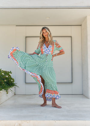 A woman standing in front of a building entrance wearing a floral and geometric patterned maxi dress with a V-neckline and billowy sleeves.