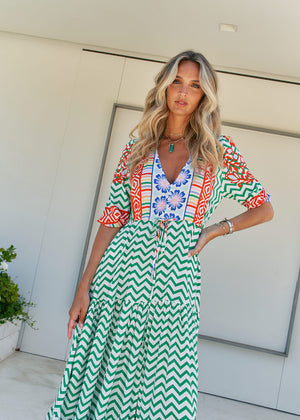 A woman standing in front of a building entrance wearing a floral and geometric patterned maxi dress with a V-neckline and billowy sleeves.