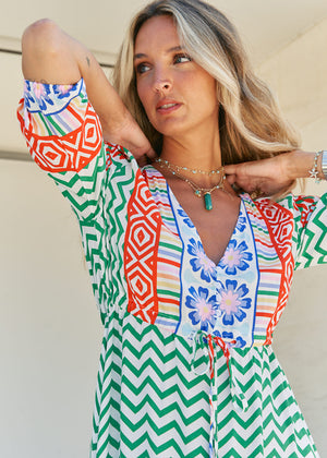 A woman standing in front of a building entrance wearing a floral and geometric patterned maxi dress with a V-neckline and billowy sleeves.
