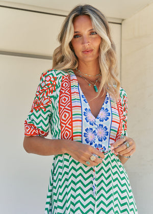A woman standing in front of a building entrance wearing a floral and geometric patterned maxi dress with a V-neckline and billowy sleeves.