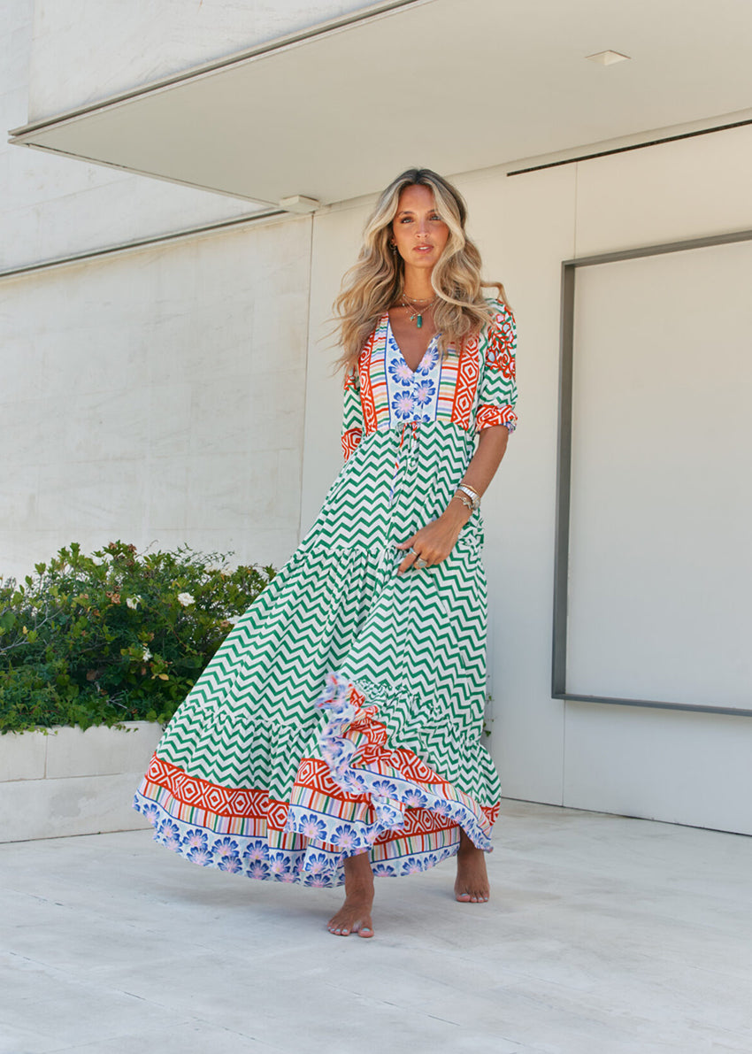 A woman standing in front of a building entrance wearing a floral and geometric patterned maxi dress with a V-neckline and billowy sleeves.