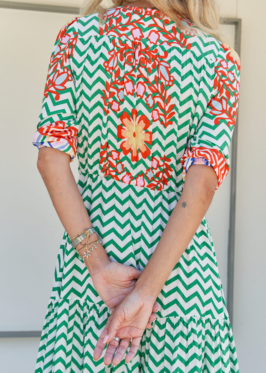 A woman standing in front of a building entrance wearing a floral and geometric patterned maxi dress with a V-neckline and billowy sleeves.