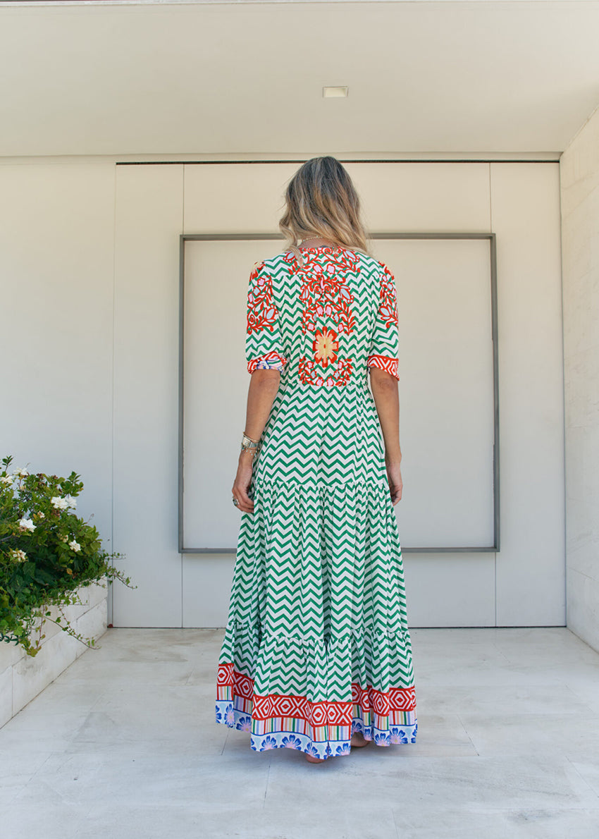 A woman standing in front of a building entrance wearing a floral and geometric patterned maxi dress with a V-neckline and billowy sleeves.