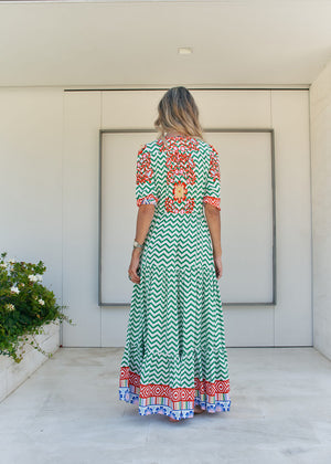 A woman standing in front of a building entrance wearing a floral and geometric patterned maxi dress with a V-neckline and billowy sleeves.