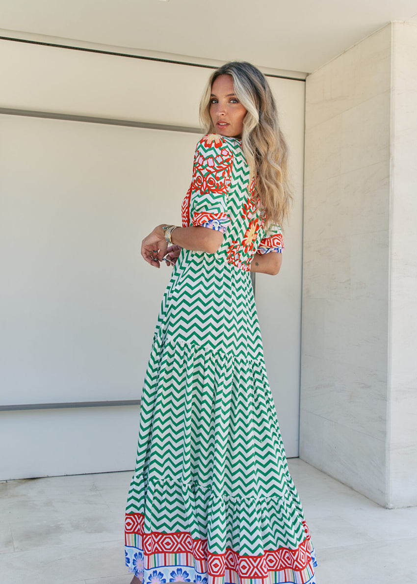 A woman standing in front of a building entrance wearing a floral and geometric patterned maxi dress with a V-neckline and billowy sleeves.