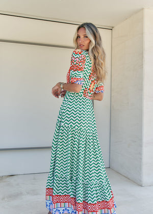 A woman standing in front of a building entrance wearing a floral and geometric patterned maxi dress with a V-neckline and billowy sleeves.