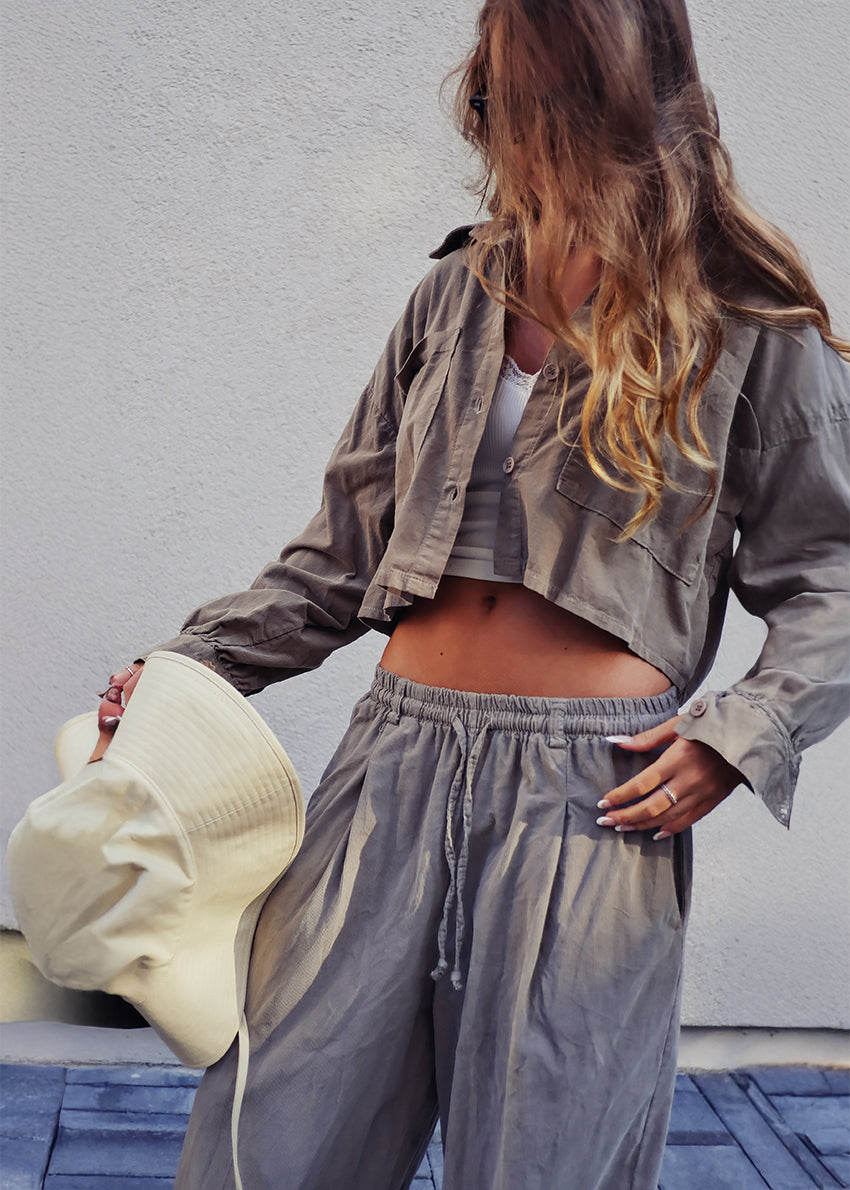 Person wearing a beige hat, light gray jacket, white crop top, and gray pants against a white wall.