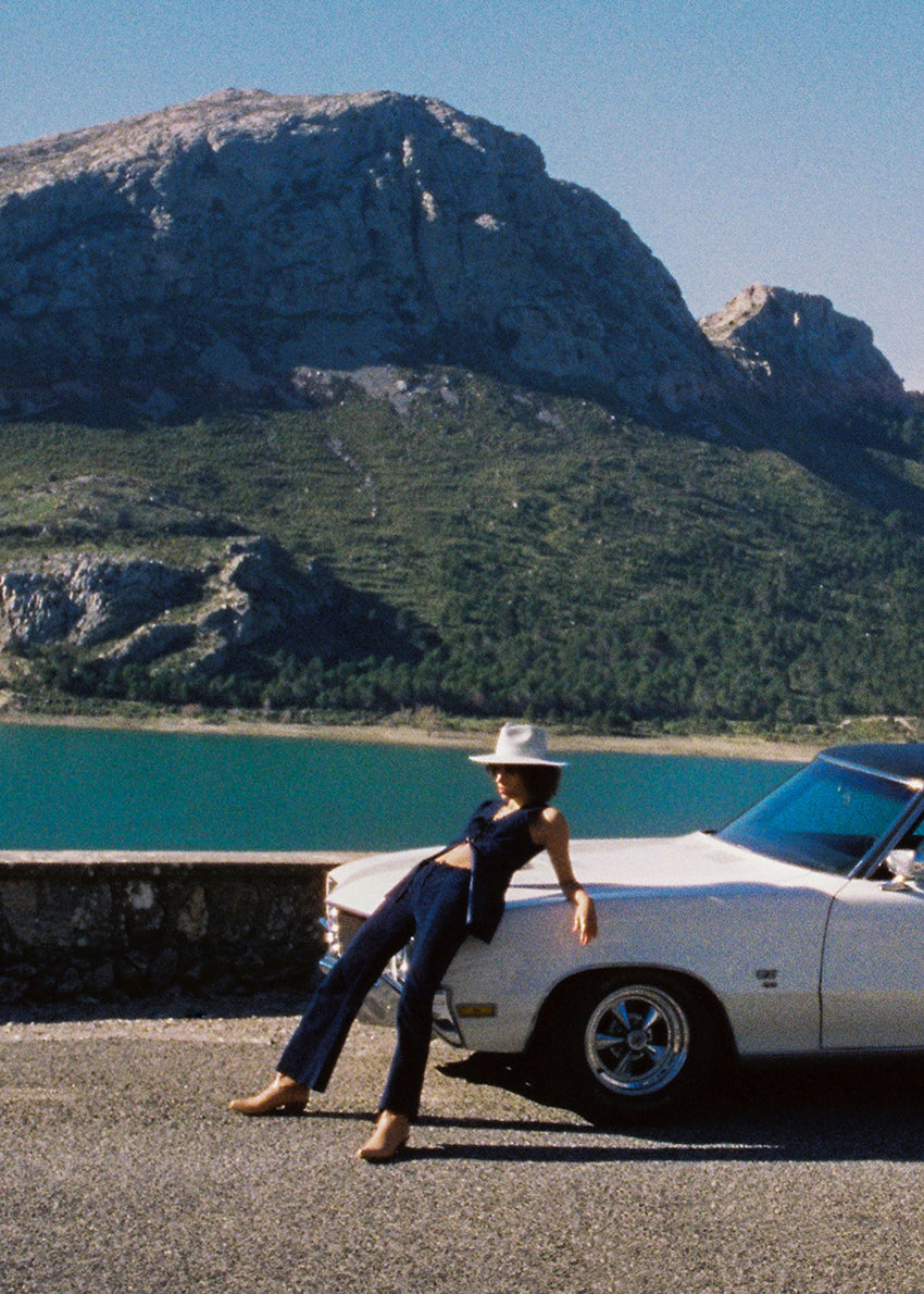 Person sitting on a classic car by a lake with mountains in the background