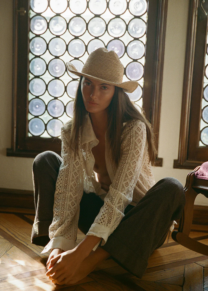Woman wearing a straw hat and lace top sitting in front of a decorative window.