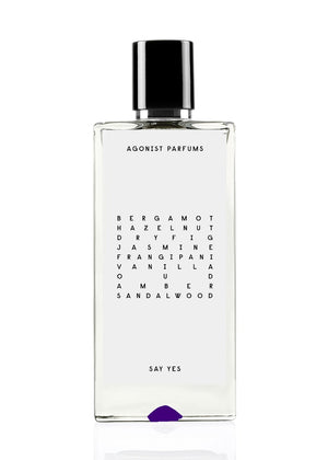 A clear glass bottle of Agonist Parfums' Say Yes Unisex Eau de Parfum with a black cap. The label lists the fragrance notes: bergamot, hazelnut, dry fig, jasmine, frangipani, vanilla, oud, amber, sandalwood.