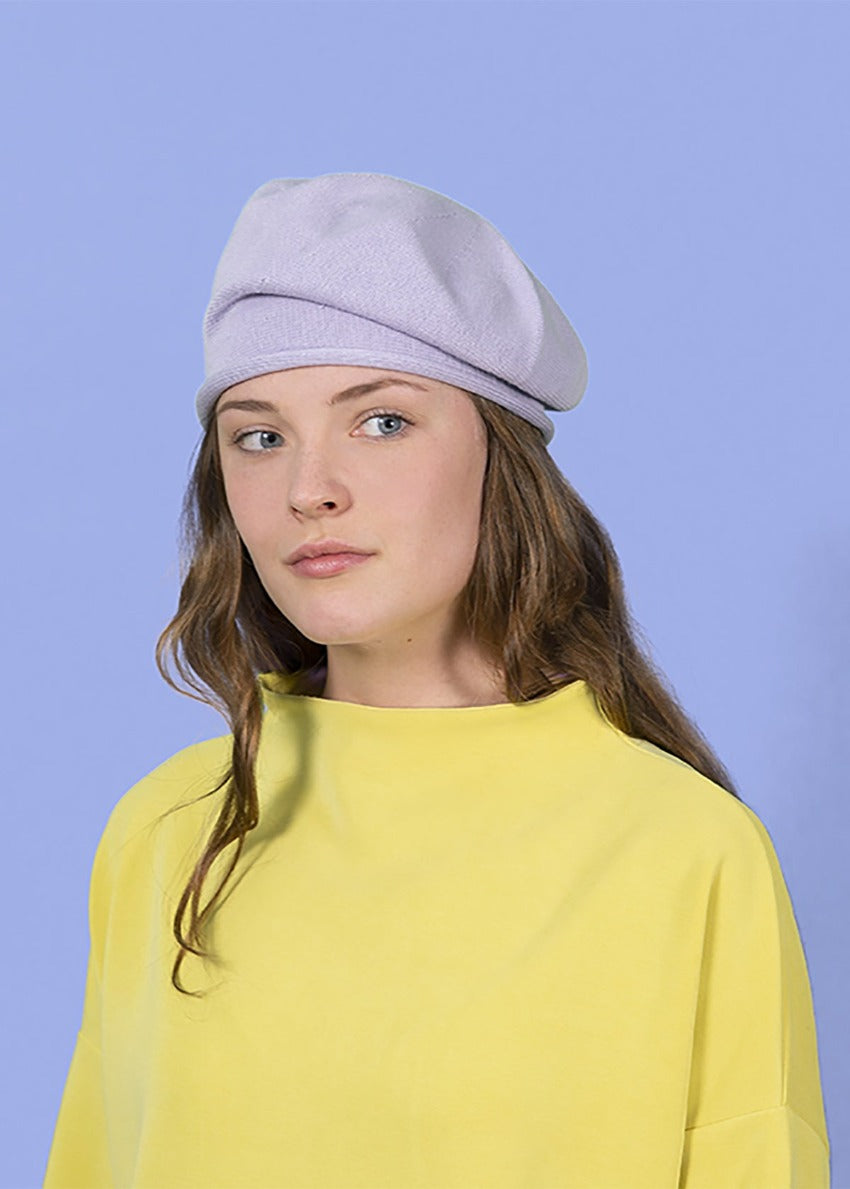 Woman wearing a beige beanie beret with a yellow sweater ob a purple background 