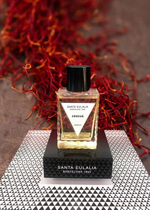 A bottle of 'Santa Eulalia' branded perfume labeled 'Crocus' with a triangular label on a reflective surface, with saffron threads in the background.