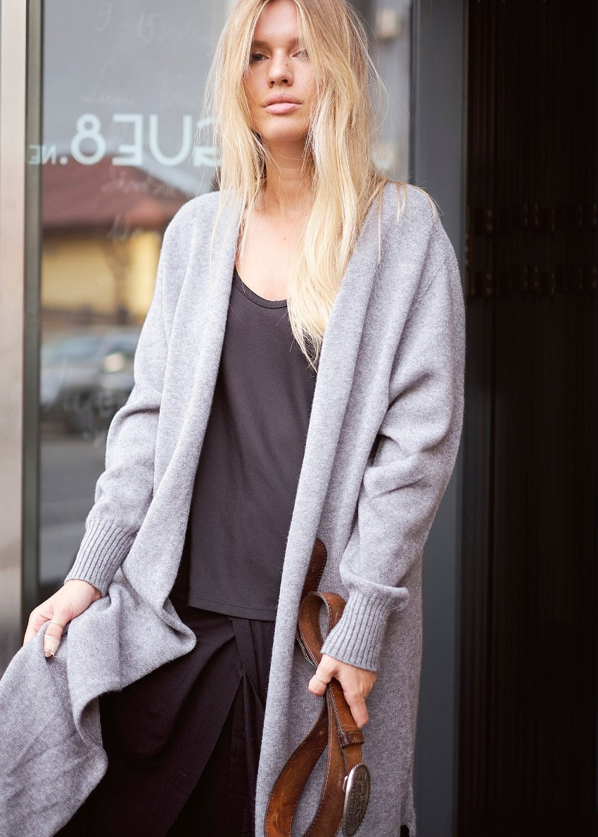 A woman standing on the sidewalk wearing a gray cardigan with long sleeves, a round neckline, and ribbed detailing. She also has a black top and is holding a brown belt.