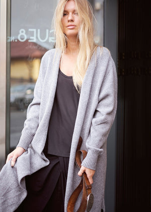 A woman standing on the sidewalk wearing a gray cardigan with long sleeves, a round neckline, and ribbed detailing. She also has a black top and is holding a brown belt.