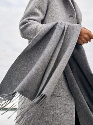 A person wearing a gray wind shawl with fringe detail, holding it with one hand against a windy backdrop.