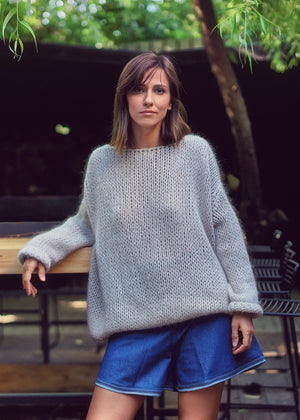 A person wearing a light grey knit cardigan with blue shorts on a nature background 