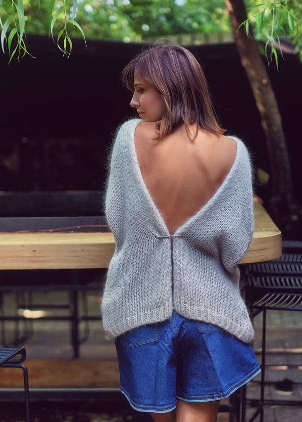 A person wearing a light grey knit cardigan with blue shorts from the back on a nature background 