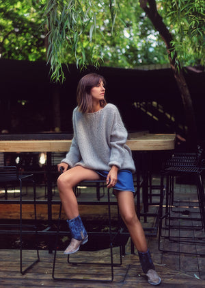 A person wearing a light grey knit cardigan with blue shorts and cowboy boots on a chair in nature