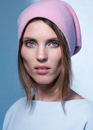 A person wearing a light pink wool beanie with a white sweater on a blue background 