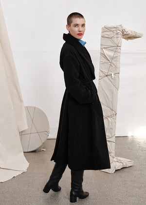 Woman wearing a black wool coat from the side with a blue shirt on a white background with different piece of furniture on the floor