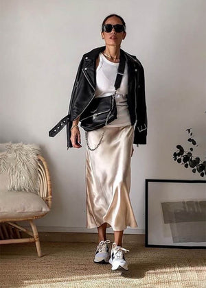 A person wearing a beige satin midi skirt with a high-rise waist and a midi-length hem, paired with a black leather jacket and white sneakers. Accessories include sunglasses and a chain shoulder bag.