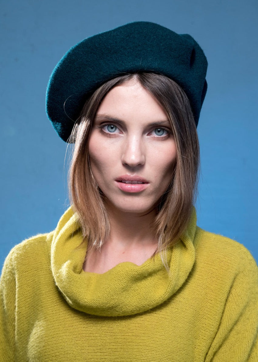 A person wearing a wool black beret with a yellow green sweater on a blue background 