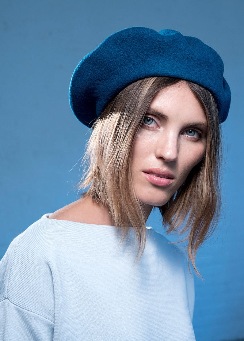 A person wearing a wool blue beret with a white sweater on a blue background 